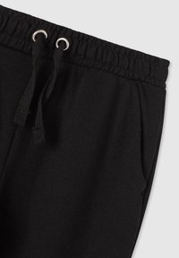 Czarna talia dresów z elastycznym paskiem i czarnymi sznurkami przewleczonymi przez metalowe oczka, z widoczną boczną kieszenią.