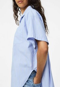 Kreklu ar īsām piedurknēm zilā un baltā svītrā, kas satur kabatu, brīvu piegriezumu un augstu-zemu hemu. Audums izskatās viegls un teksturēts.