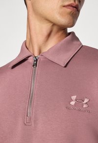 Rózsaszín cipzáros póló, gallérral, texturált anyagból készült, bal mellen hímezett Under Armour logóval.