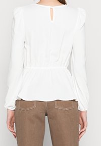 Blusa bianca con maniche a palloncino, con vita arricciata e dettaglio a occhiello sul retro, abbinata a pantaloni marroni a trama con tasche posteriori.