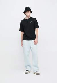 Svart T-skjorte med en liten hvit logo, lyseblå vidbukser, svart bøttehatt og grå joggesko med svarte og hvite detaljer.