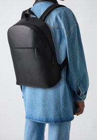 Osoba v černém koženém batohu s přední kapsou na zip, v modré džínové bundě a džínech, na hladkém pozadí.