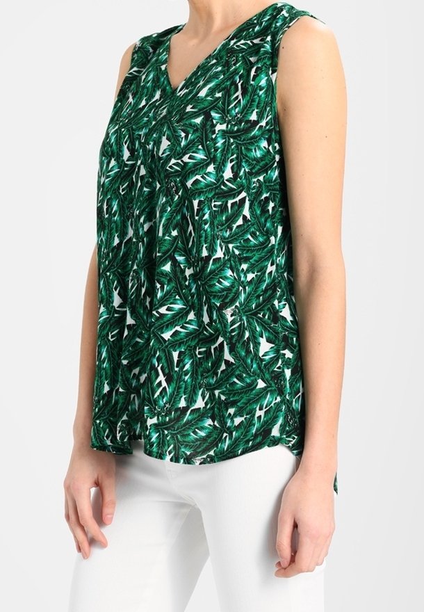 Blouse sans manches à motif feuilles tropicales vertes et blanches portée avec un pantalon blanc sur une personne debout sur un fond uni.