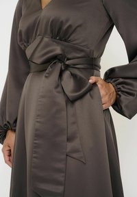 Braunes Satin-Kleid mit taillierter Silhouette, das mit einer Schleifenapplikation, langen Puffärmeln und einer glatten, glänzenden Textur versehen ist.