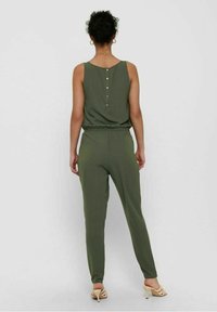 Olijfgroene jumpsuit met een mouwloos ontwerp, elastische tailleband en knoopdetails aan de achterkant. Gladde stof en taps toelopende benen.