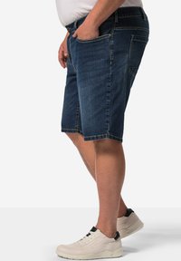 Shorts en denim bleu foncé avec une coupe droite, cinq poches et des coutures visibles. Portés avec une chemise blanche et des sneakers de couleur claire.