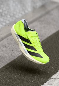 Zapatilla de correr de un brillante color amarillo neón con rayas negras, suela blanca y cordones, suspendida sobre un fondo de asfalto y pavimento.