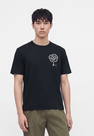 Homme portant un t-shirt noir avec un motif de fleur blanche sur la poitrine et un pantalon vert olive, debout devant un fond clair uni.
