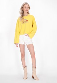 Žlutá zkrácená mikina s květinovou grafikou, spárovaná s bílými distressed šortkami a béžovými kotníkovými botami s texturovaným povrchem.