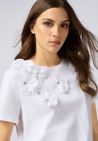 Witte katoenen T-shirt met een getextureerd bloem appliqué ontwerp en heldere rhinestones langs de halslijn. Korte mouwen en een ronde hals.