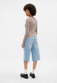 Un top lung cu maneci, de culoare maro si textură ribbed, asortat cu pantaloni scurți lejeri din denim albastru deschis și pantofi platform negri, având un tiv brut. Sosetele albe sunt de asemenea vizibile.