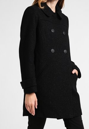 Femme portant un manteau en laine noir longueur genou avec des boutons à double boutonnage, des poches latérales et des boutons aux poignets, posant avec une main dans la poche.