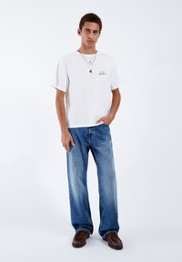 White cotton t-shirt with a small black logo, paired with high-waisted faded blue jeans and dark brown leather shoes.