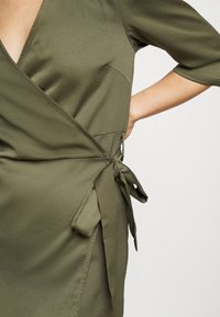 Femme portant une robe portefeuille vert olive à manches mi-longues, nouée en un nœud à la taille, la main posée sur la hanche.