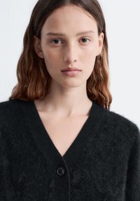 Schwarzer, flauschiger Cardigan mit V-Ausschnitt, versehen mit einem schwarzen Knopf in der Mitte. Die Textur wirkt weich und gestrickt.