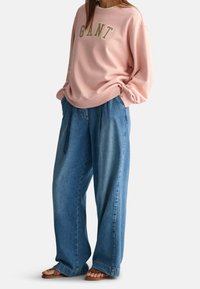 Sweat-shirt rose avec le logo "GANT" en lettres dorées, associé à un pantalon en denim bleu ample. Sandales marron complètent la tenue.