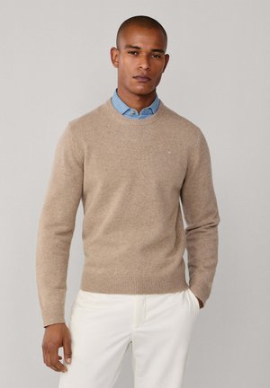 Suéter beige de cuello redondo hecho de un material suave y texturizado, usado sobre una camisa de cuello azul claro, con puños y dobladillo de canalé.