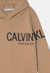 Tan hoodie i mjukt material, med fet svart text "CALVIN KLEIN" och "ETABLERAD 1978" tryckt på framsidan.