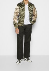 Quiltad bomberjacka i olivgrön med gulddetaljer, har ribbad krage och manschetter, matchad med mörka byxor och vita/gröna sneakers.