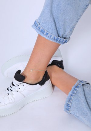 Weißer Sneaker mit schwarzen Akzenten, ausgestattet mit einer strukturierten Sohle und Schnürdesign. Getragen mit hochgekrempelten, hellblauen Jeans. Silbernes Fußkettchen sichtbar.