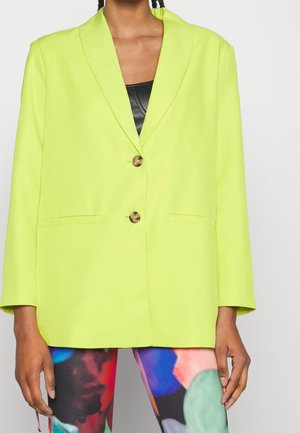 Kobieta ubrana w limonkową marynarkę na dwa guziki, czarny top i legginsy z wielokolorowym abstrakcyjnym wzorem, stojąca na białym tle.