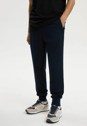 Mężczyzna w czarnych dresach, beżowo-granatowych sneakersach, stojący z jedną ręką w kieszeni na jednolitym jasnym tle.