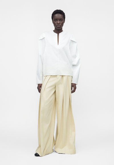 Femme aux cheveux courts porte un haut blanc à manches longues avec des détails aux épaules et un pantalon beige large plissé, debout sur fond blanc.