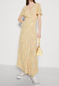 Robe portefeuille florale jaune avec des manches courtes et un décolleté en V. Présente un design fluide, un tissu superposé, portée avec des baskets blanches et un sac jaune.