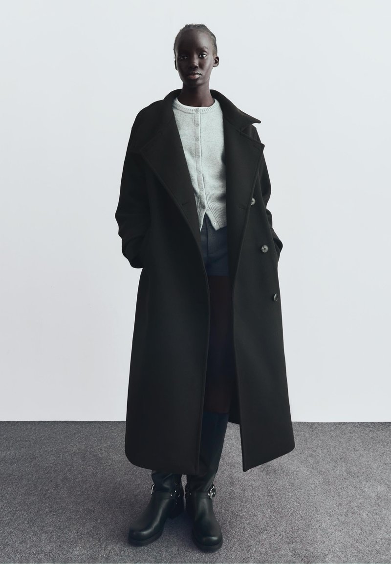 Lång person som bär en lång svart kappa, ljusgrå knäppt cardigan, mörka shorts och svarta knähöga stövlar som står på grå matta mot vit vägg.