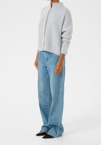 Hellgraue Strickjacke mit hohem Kragen, Knopfverschluss und weitem Schnitt, kombiniert mit hellblauen Jeans mit weitem Bein und schwarzen spitzen Schuhen.