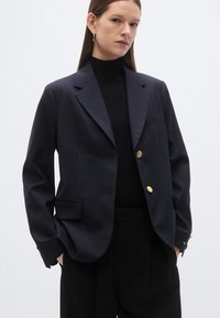 Marineblauw colbert in een aansluitend ontwerp met ingesneden revers, gouden knopen, twee voorzakken en een enkele borstzak, gecombineerd met een zwart topje.
