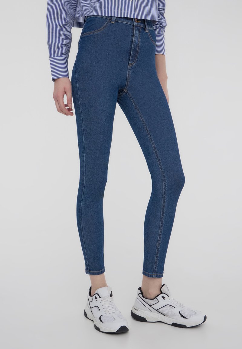 Jeans in denim blu a vita alta con una vestibilità slim, caratterizzati da una texture liscia e cuciture di contrasto, abbinati a sneakers bianche.