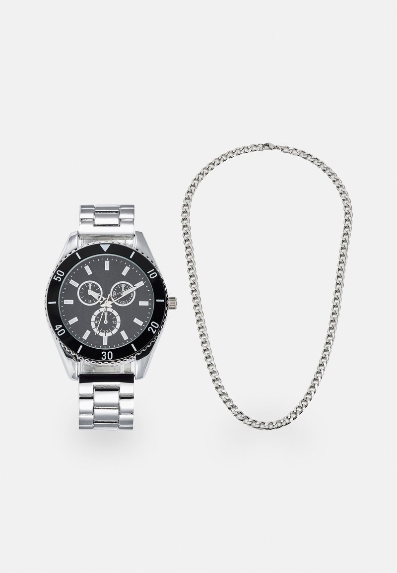 Montre en métal argenté avec un cadran noir, fonctionnalités chronographe, bracelet maillons, et un collier en chaîne argenté placé à côté.