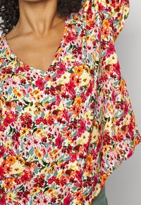 Blouse florale en tissu léger présentant un mélange vibrant de fleurs rouges, roses et orange sur un fond blanc. Col en V avec boutons.