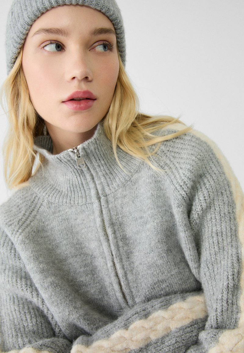 Maglione grigio a maglia con colletto alto, apertura con mezza zip sul davanti, motivo testurizzato e dettagli color crema; abbinato a un berretto grigio coordinato.