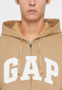 Béžová mikina s kapucňou na zips vyrobená z jemného materiálu, s bielym logom "GAP". Obsahuje šnúrky a veľkú kapucňu so hladkým uzáverom na zips.