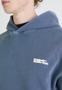 Minkšta, mėlyna medvilninė gobtuvinė palaidinė su laisvu fasonu. Dekoruota balta logotipų tekstu ant krūtinės ir toniniais siūlais. Lygi tekstūra.