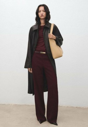 Woman wearing a long black coat over a burgundy top and pants, holding a large beige shoulder bag, standing against a plain light wall.