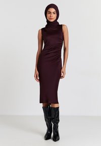 Abito senza maniche color marrone con collo alto e dettagli arricciati sui lati; abbinato a stivali lucidi neri al ginocchio. Tessuto liscio e elasticizzato.