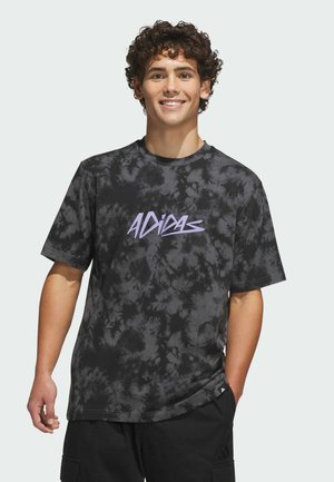 Młody mężczyzna uśmiecha się, nosi czarno-szarą koszulkę Adidas tie-dye z fioletowym logo, czarne spodnie, stoi z jedną ręką w kieszeni.