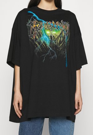 Person wearing oversized black T-shirt with neon green and blue lightning and skull graphic and stylized text on front, paired with blue jeans.