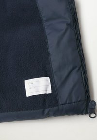 Vnitřek námořnické fleecové bundy ukazuje část zipu a bílou etiketu s políčky pro jméno a telefonní číslo.