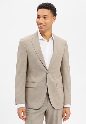Man draagt een licht beige pak met een wit overhemd, glimlachend en zijwaarts kijkend tegen een eenvoudige witte achtergrond.