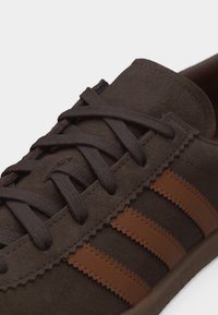 Κοντινή λήψη σκούρου καφέ σουέτ αθλητικού παπουτσιού με κορδόνια στο ίδιο χρώμα και τρεις ανοιχτόχρωμες δερμάτινες ρίγες με ζιγκ-ζαγκ άκρες στο πλάι.