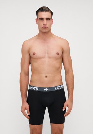 Boxer briefs noirs avec une ceinture élastique grise arborant le logo "Lacoste", texture lisse et coupe ajustée.