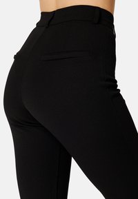 Nahaufnahme einer Person, die eng anliegende schwarze Hose mit hoher Taille und zwei aufgesetzten Gesäßtaschen trägt, vor einem schlichten Hintergrund.