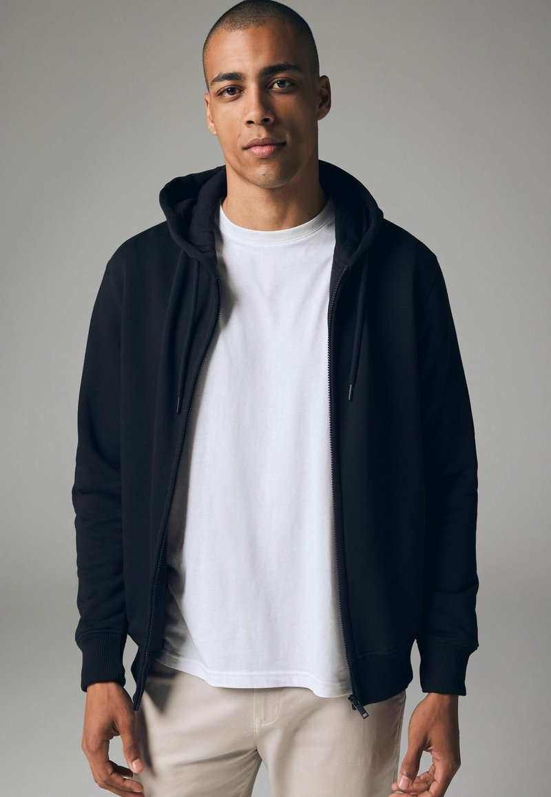 Haut de sweatshirt noir zippé avec capuche à cordon, porté sur un t-shirt blanc, avec des poignets côtelés et une texture lisse. Design simpliste.