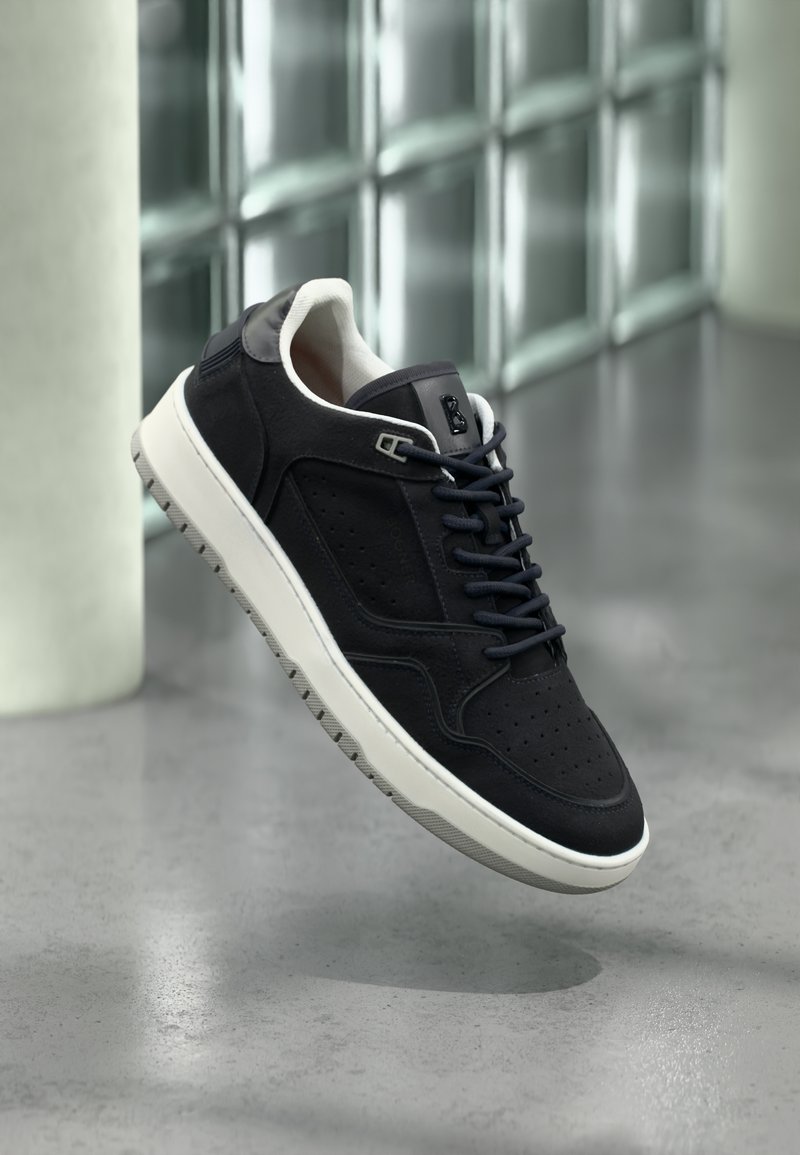 Μαύρο αθλητικό παπούτσι με λευκή σόλα και μαύρα κορδόνια που αιωρείται πάνω από γκρίζο τσιμεντένιο πάτωμα, με τοίχο από γυάλινα τουβλάκια στο παρασκήνιο.