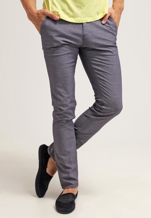 Mężczyzna w szarych dopasowanych spodniach, z rękami w kieszeniach, ubrany w czarne buty wsuwane, stojący z jedną nogą założoną na drugą.