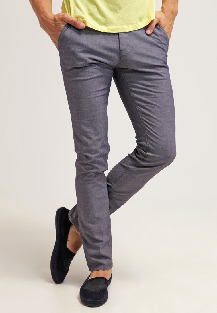 Homme portant un pantalon gris coupe ajustée, mains dans les poches et chaussures noires sans lacets, debout avec une jambe croisée sur l'autre.